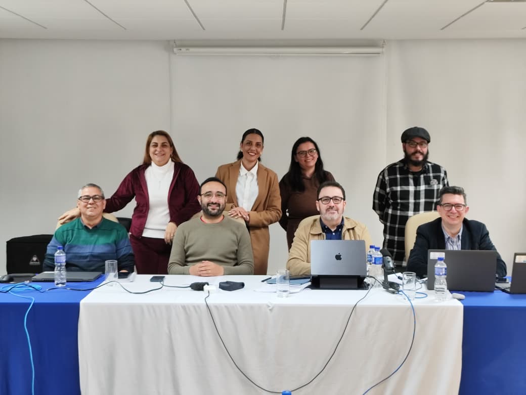 Imagem de oito pessoas: quatro sentadas e quatro em pé. Na frente da imagem, tem uma mesa azul com dois computadores. Eles estão em um ambiente de trabalho.